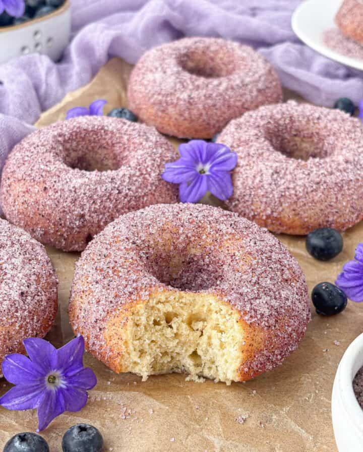 Baked doughnuts coated in purple sugar, one with a bite taken out, surrounded by blueberries and purple flowers on a wooden surface.