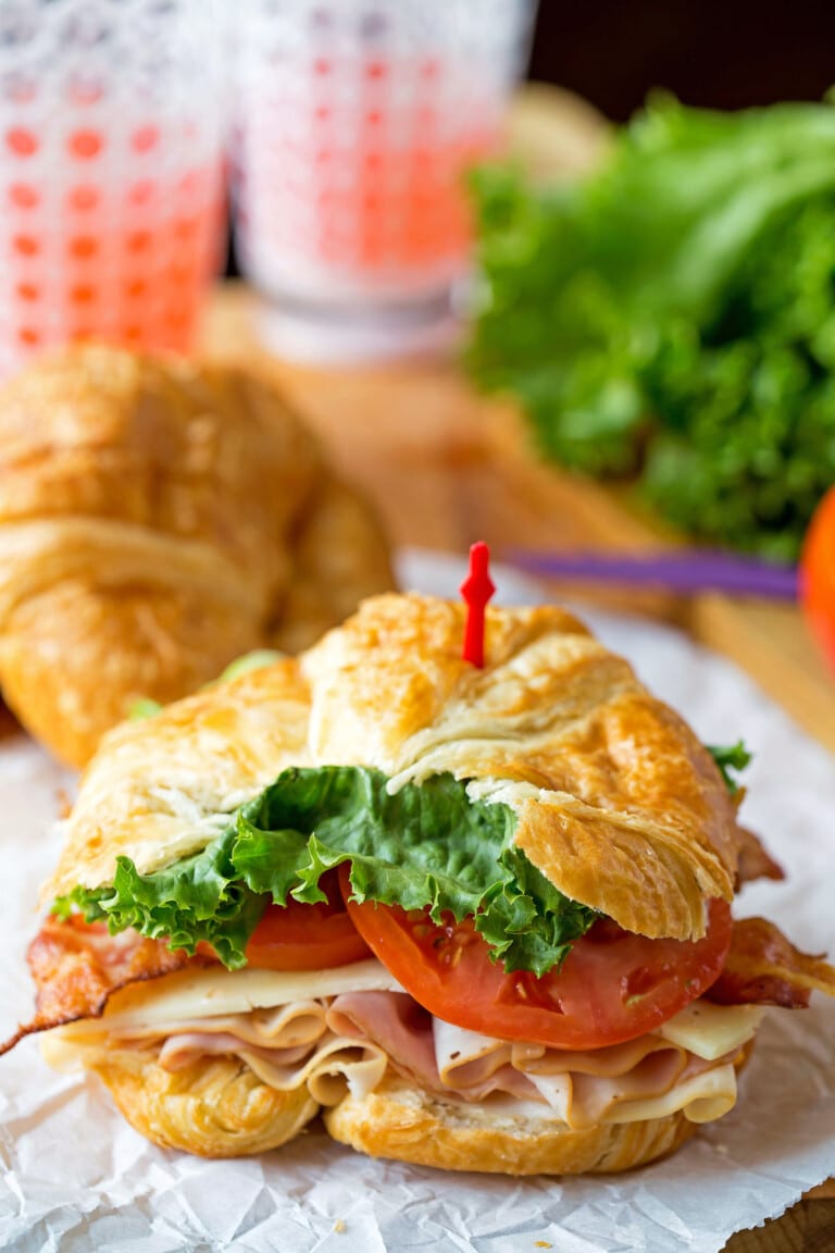 A croissant sandwich filled with turkey, bacon, cheese, lettuce, and tomato, secured with a red cocktail stick, sits on greaseproof paper with another croissant and drinks in the background.