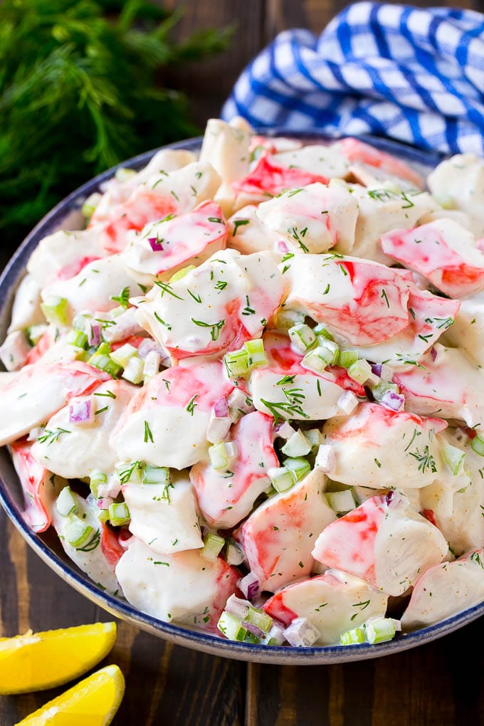 A bowl of creamy crab salad with chunks of imitation crab, diced celery, red onion, and fresh dill, served on a wooden table with lemon wedges nearby.