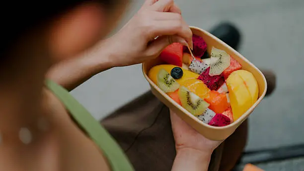 A person holding a container filled with assorted sliced fruits including kiwi, mango, melon, dragon fruit, blueberry, and orange.
