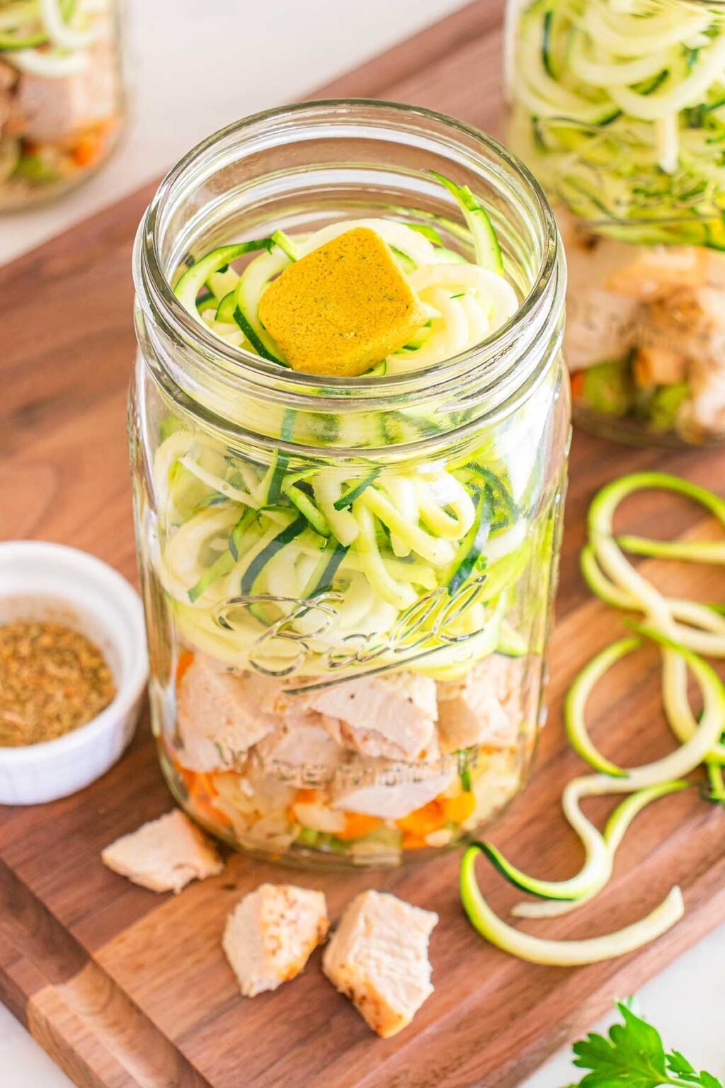 A jam jar filled with cubed chicken, spiralised courgette, diced carrots, and a stock cube, placed on a wooden board with scattered courgette spirals.