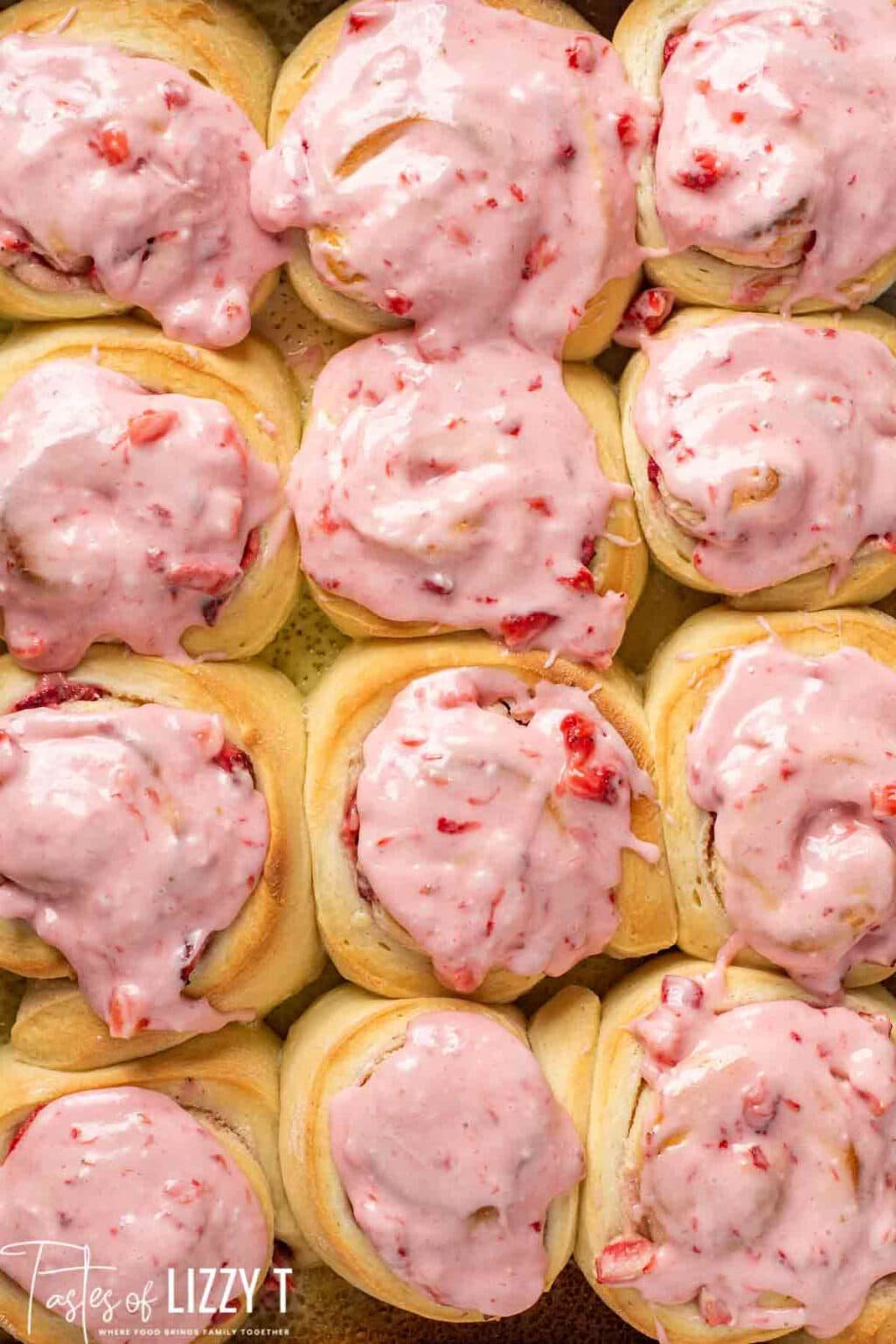 A close-up of twelve baked rolls topped with thick, pink strawberry icing arranged in a grid pattern.