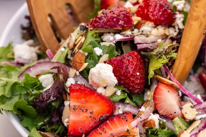 Mixed green salad with sliced strawberries, red onions, crumbled cheese, almond slices, and poppy seeds, being tossed with wooden salad servers.