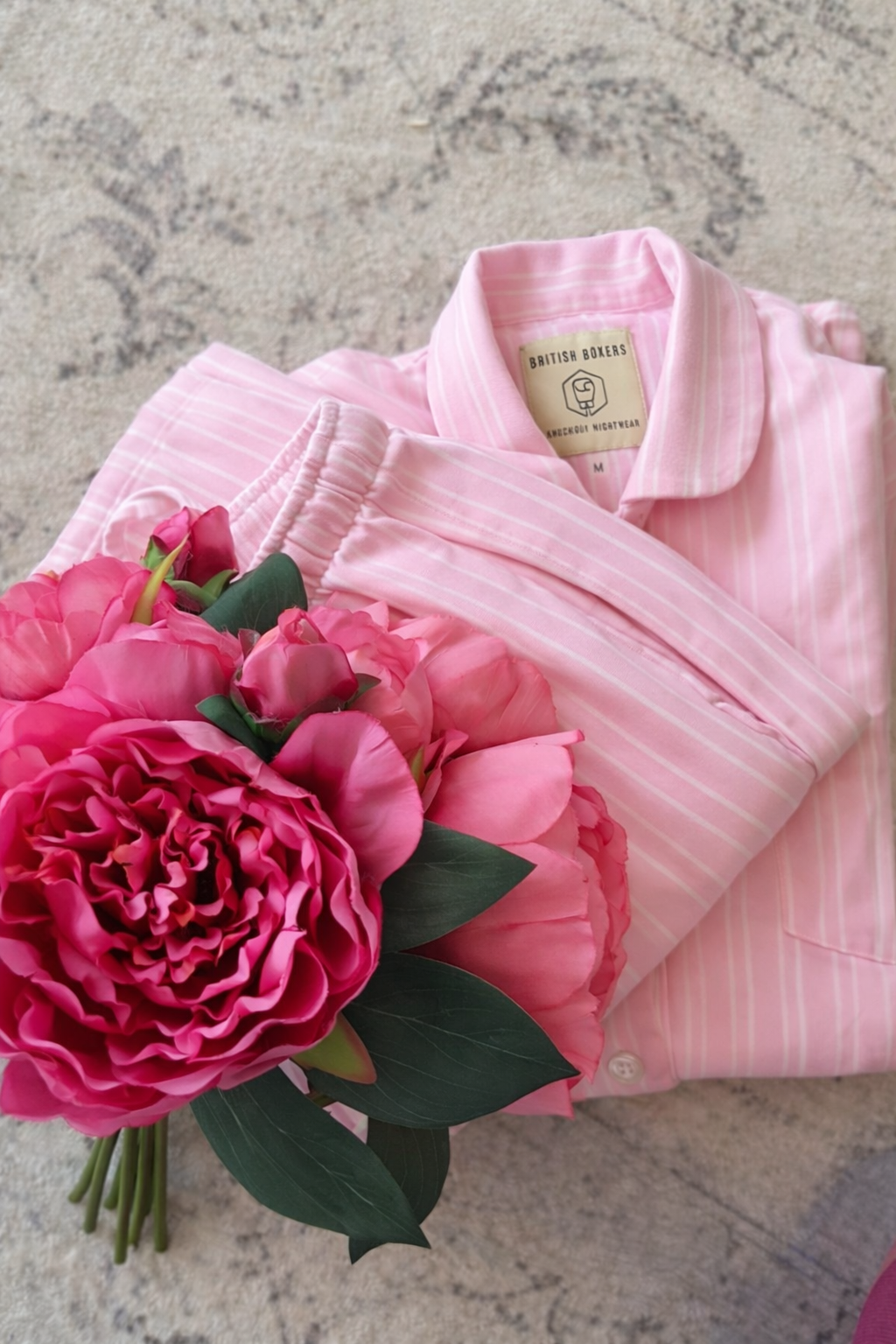 A folded Pink Stripe Pyjamas shirt from British Boxers, topped with a bouquet of bright pink flowers, rests elegantly on a patterned surface.
