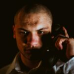 A person with a serious expression holds a black telephone receiver to their ear, partially lit against a dark background-capturing the tension often faced by an FCA Whistleblower considering contacting an FCA Whistleblower Solicitor.