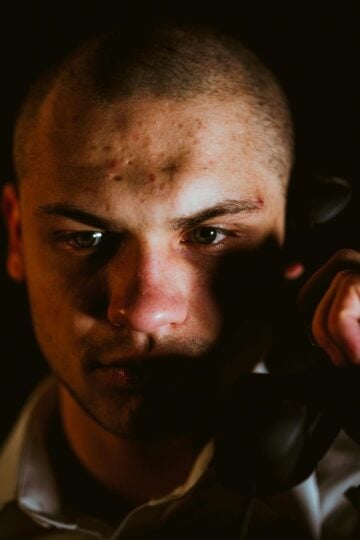 A person with a serious expression holds a black telephone receiver to their ear, partially lit against a dark background—capturing the tension often faced by an FCA Whistleblower considering contacting an FCA Whistleblower Solicitor.
