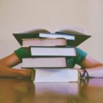 A person sits at a table with their head and arms behind a tall stack of books, with one book open on top of the pile-looking for revision tips to improve your marks.