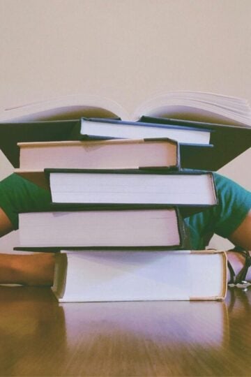 A person sits at a table with their head and arms behind a tall stack of books, with one book open on top of the pile—looking for revision tips to improve your marks.