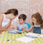 An adult and two preschoolers sit at a green polka dot table, painting with brushes and watercolours in a classroom with wall art, enjoying predictable routines that help them feel secure.