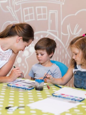 An adult and two preschoolers sit at a green polka dot table, painting with brushes and watercolours in a classroom with wall art, enjoying predictable routines that help them feel secure.