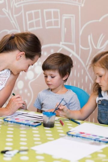 An adult and two preschoolers sit at a green polka dot table, painting with brushes and watercolours in a classroom with wall art, enjoying predictable routines that help them feel secure.