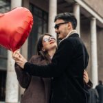 A couple embraces outdoors, with the woman holding a red heart-shaped balloon. The man's coat and glasses create a confident impression-perfect inspiration for a first date outfit men will want to try in 2026, set against columned architecture.