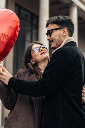 A couple embraces outdoors, with the woman holding a red heart-shaped balloon. The man’s coat and glasses create a confident impression—perfect inspiration for a first date outfit men will want to try in 2026, set against columned architecture.