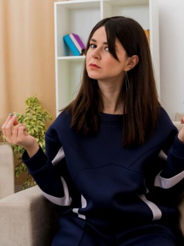 A woman with long dark hair in a navy sweatshirt sits indoors on a beige chair, holding her hands up with an indifferent or questioning expression-perhaps searching for ways to beat brain fog and reset clarity.
