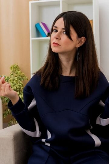 A woman with long dark hair in a navy sweatshirt sits indoors on a beige chair, holding her hands up with an indifferent or questioning expression—perhaps searching for ways to beat brain fog and reset clarity.