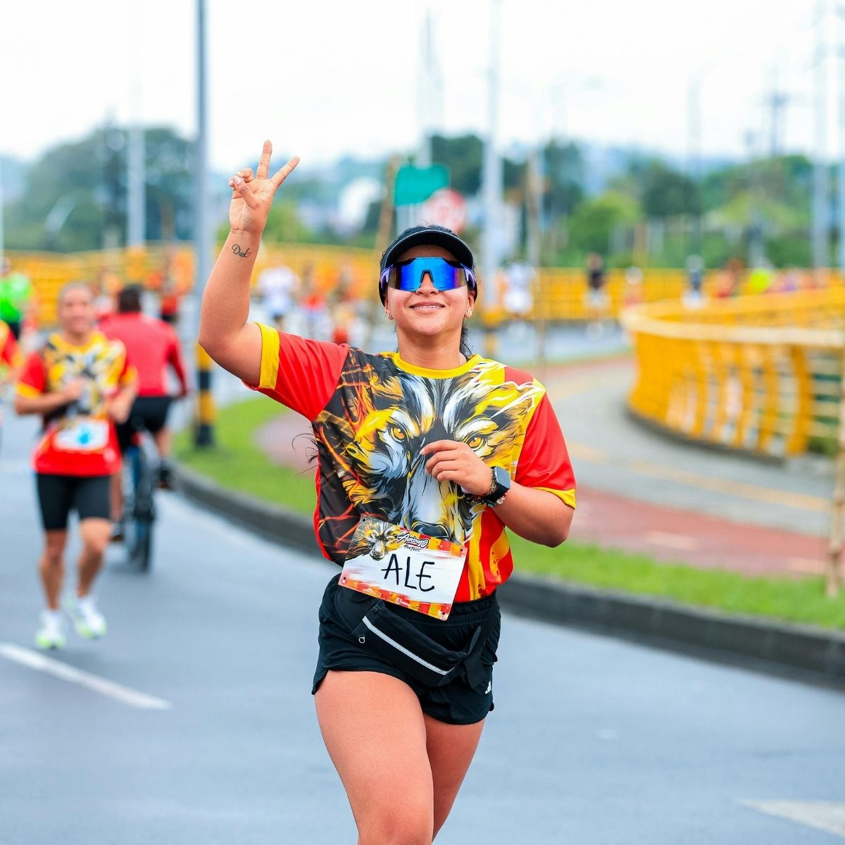 Runner wearing sunglasses and a lion-print shirt raises a peace sign whilst participating in a road race; other runners are visible in the background.