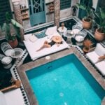 A woman in a swimming costume and sunhat lounges on a large outdoor bed beside a turquoise pool-a must-have retreat for pool owners-set in a courtyard with white furniture and potted plants.