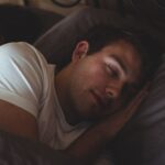 A person wearing a white shirt is lying in bed, sleeping on their side with their head resting on a pillow, enjoying restful sleep.