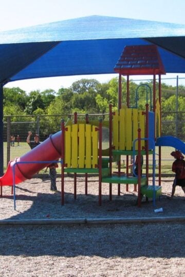 Children enjoy outdoor adventures on a colourful playground structure with a slide under a blue shade canopy, with a sports pitch and trees in the background—perfect for family fun and kids’ activities.