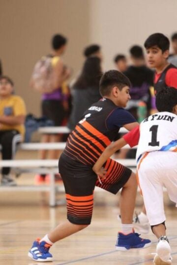 Two boys play basketball indoors, showcasing family sport as others watch and sit on benches in the background. The player in black and orange guards the player in white and red with the ball, highlighting fun activities for everyone.