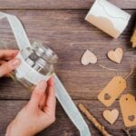 Hands wrapping lace ribbon around a small glass jar on a wooden table, surrounded by wooden hearts, kraft paper tags, a ribbon roll, and a stick-capturing the crafting joy of creative DIY ideas at home.