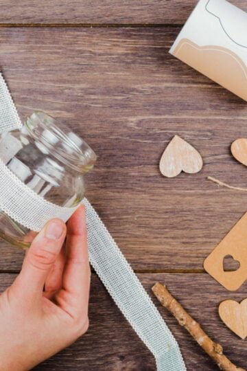 Hands wrapping lace ribbon around a small glass jar on a wooden table, surrounded by wooden hearts, kraft paper tags, a ribbon roll, and a stick—capturing the crafting joy of creative DIY ideas at home.