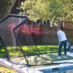 A person is practising golf swings on a back garden turf mat, aiming at a large black net set up near a swimming pool to master golf at home and improve golfing skills.