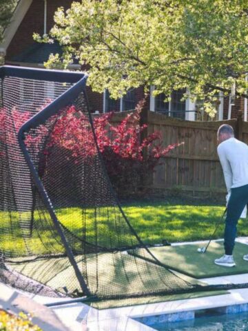 A person is practising golf swings on a back garden turf mat, aiming at a large black net set up near a swimming pool to master golf at home and improve golfing skills.