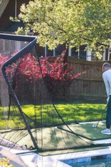 A person is practising golf swings on a back garden turf mat, aiming at a large black net set up near a swimming pool to master golf at home and improve golfing skills.