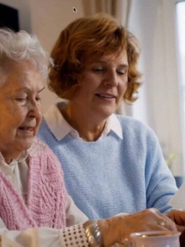 Two women sit together at a table, looking at papers. One is an elderly senior with white hair and a pink waistcoat, the other has brown hair and wears a light blue jumper. They discuss ways to live independently at home.