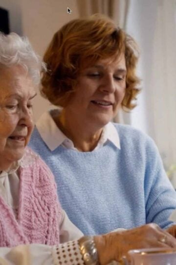 Two women sit together at a table, looking at papers. One is an elderly senior with white hair and a pink waistcoat, the other has brown hair and wears a light blue jumper. They discuss ways to live independently at home.