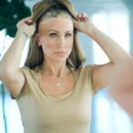 A woman stands in front of a mirror, lifting a brown wig off her head to reveal a shaven scalp underneath-an empowering step towards restoring hair confidence and embracing expert hair advice.