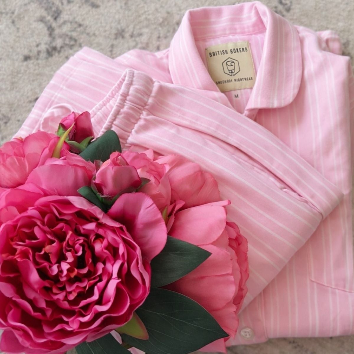 A folded set of Pink Stripe Pyjamas with a British Boxers label, next to a bouquet of pink artificial flowers on a light surface—perfect for your next Pyjamas Review.