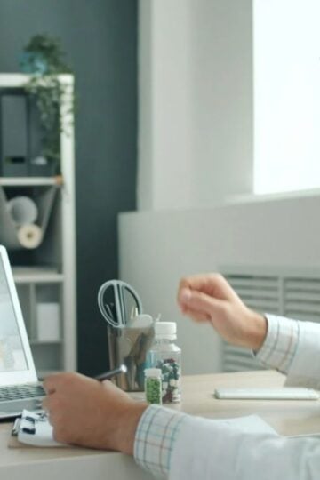 A doctor in a white coat and glasses sits at a desk, offering GP advice to busy families via video call on a laptop, with medicine bottles and office supplies nearby.