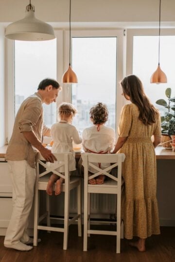 Two adults and two children stand and sit at a kitchen worktop, facing a window. The kitchen is bright with white cupboards and pendant lights.
