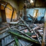 Interior of a damaged, abandoned building with collapsed wooden beams, renovation debris scattered on the floor, and partially destroyed walls revealing exposed brickwork.
