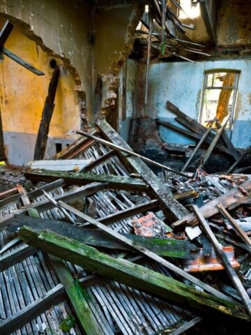 Interior of a damaged, abandoned building with collapsed wooden beams, renovation debris scattered on the floor, and partially destroyed walls revealing exposed brickwork.