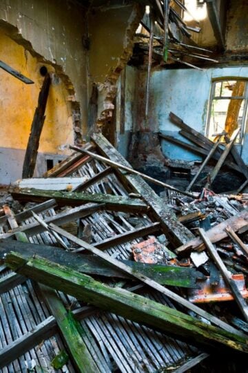 Interior of a damaged, abandoned building with collapsed wooden beams, renovation debris scattered on the floor, and partially destroyed walls revealing exposed brickwork.