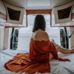 A woman with shoulder-length hair sits on a bed in a van, draped in a rust-coloured cloth, gazing out the open back doors towards a beach and sky-embracing the freedom of van life over traditional housing.
