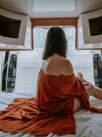 A woman with shoulder-length hair sits on a bed in a van, draped in a rust-coloured cloth, gazing out the open back doors towards a beach and sky-embracing the freedom of van life over traditional housing.