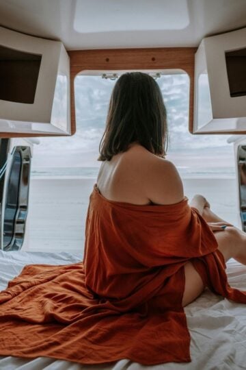 A woman with shoulder-length hair sits on a bed in a van, draped in a rust-coloured cloth, gazing out the open back doors towards a beach and sky—embracing the freedom of van life over traditional housing.