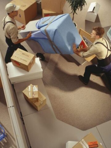 Two removal men in uniform carry a large padded item through an office filled with boxes, packing materials, and disassembled furniture-showcasing the efficiency of stress-free removal services for home removals Singapore.