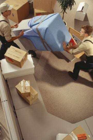 Two removal men in uniform carry a large padded item through an office filled with boxes, packing materials, and disassembled furniture—showcasing the efficiency of stress-free removal services for home removals Singapore.