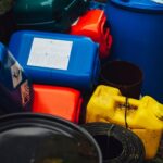 Colourful plastic containers, including blue, red, and yellow jerrycans, are stacked outdoors alongside black cylindrical objects-ideal as a COSHH container solution for safe chemical storage.