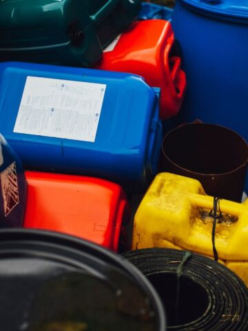 Colourful plastic containers, including blue, red, and yellow jerrycans, are stacked outdoors alongside black cylindrical objects-ideal as a COSHH container solution for safe chemical storage.