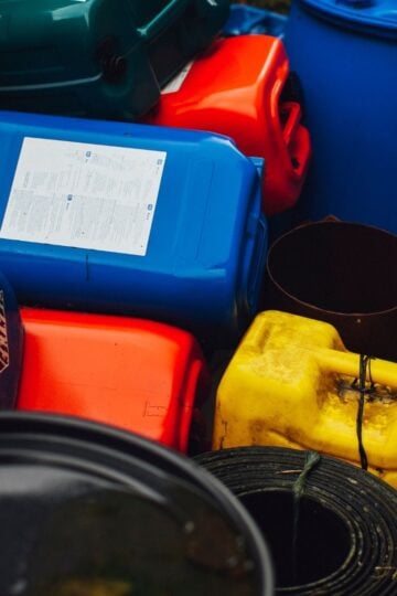 Colourful plastic containers, including blue, red, and yellow jerrycans, are stacked outdoors alongside black cylindrical objects—ideal as a COSHH container solution for safe chemical storage.
