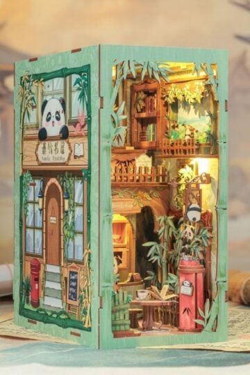 A detailed miniature diorama of a two-storey panda-themed café crafted like wooden book nook kits, featuring bamboo décor, warm lighting, and intricate furniture, with structural panels that align seamlessly against a blurred floral landscape background.
