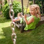 A young child in a green dress smiles whilst swinging on a swing in a sunny children's garden, enjoying outdoor play.
