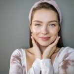 A woman with light eyes and a hairband smiles softly, resting her hands on her cheeks, subtly highlighting the natural beauty of ageing skin against a plain grey background.