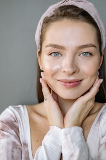 A woman with light eyes and a hairband smiles softly, resting her hands on her cheeks, subtly highlighting the natural beauty of ageing skin against a plain grey background.